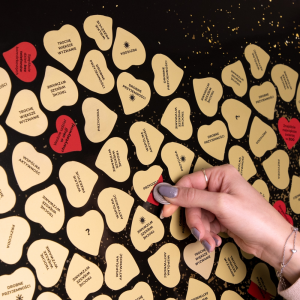 Zbliżenie na plakat Zdrapka Przeżyć dla Pary, kobieta zdrapuje kolejne pola. Na zdjęciu widać przykładowe przeżycia: Weźcie udział w akcji charytatywnej, Spędźcie dzień bez telefonów komórkowych